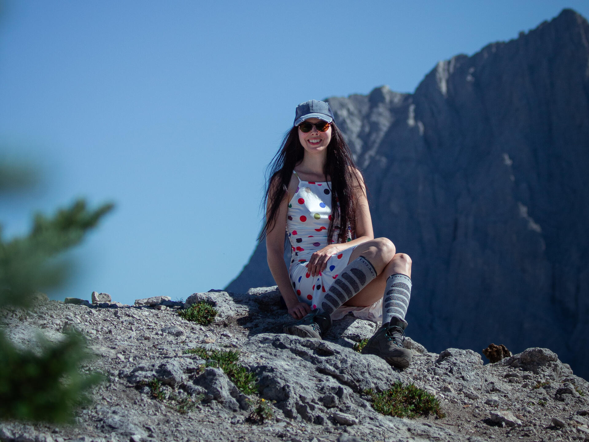 A smiling young woman with long brown hair, wearing a polkadot dress, hiking boots, and a ball cap sits on a rock in the alpine